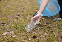 A woman hands in gloves collects and puts used plastic bottle into a blue trash bag. A volunteer cleans up the park on a sunny bright day. Clearing, pollution, ecology and plastic concept.