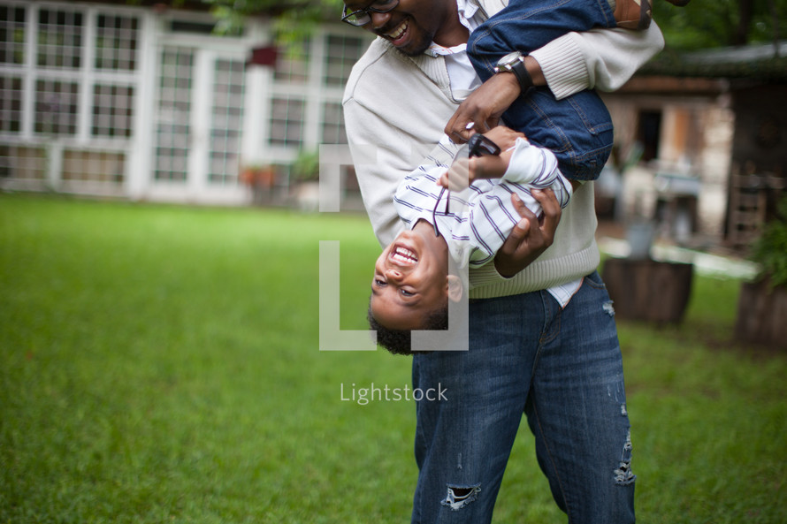 A father holding and playing with his young son in the back yard.