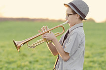 Boy in a hat playing trumpet in a grassy meadow.