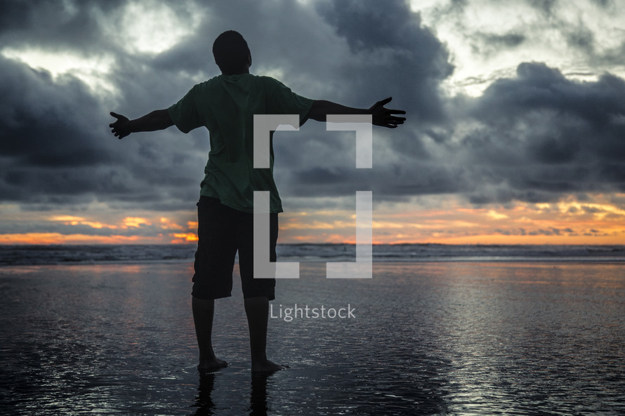 a man standing on a beach on wet sand with open arms at sunset 