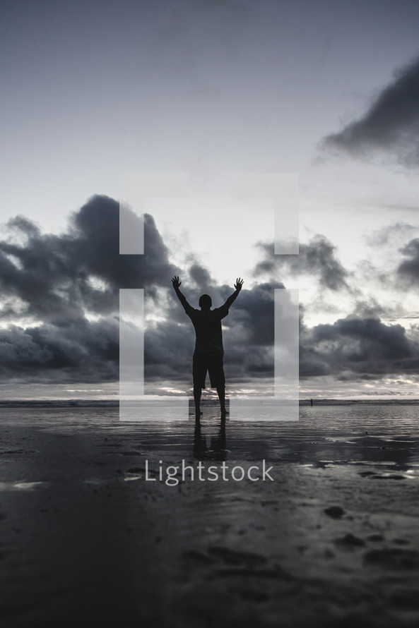 a man standing on a beach on wet sand with hands raised at sunset 