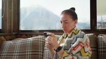 Pretty woman sitting on the cafe terrace on the background of winter mountain landscape, using a smartphone. Woman tourist uses internet on smartphone on restaurant in mountain. Amazing mountains view.