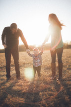 Man and woman holding hands of toddler child in grassy field at sunset.