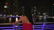 Young beautiful woman stand in evening city against multi-storey builds in chic evening red dress and leaning on railing with hand admire view of river and passing away ships. Happy and freedom life.