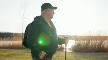 Active handsome elderly man with nordic walking sticks walks forward through along the lake at sunset. Senior man is engaged in scandinavian Walking with a backpack. Healthy lifestyle concept.