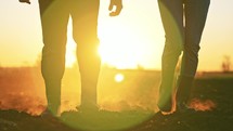 Close up people feet are walking on black earth soil on agricultural land in rubber boots in nature on field in summer at sunset and dust rises in sun. Couple of rural farmers in family business.