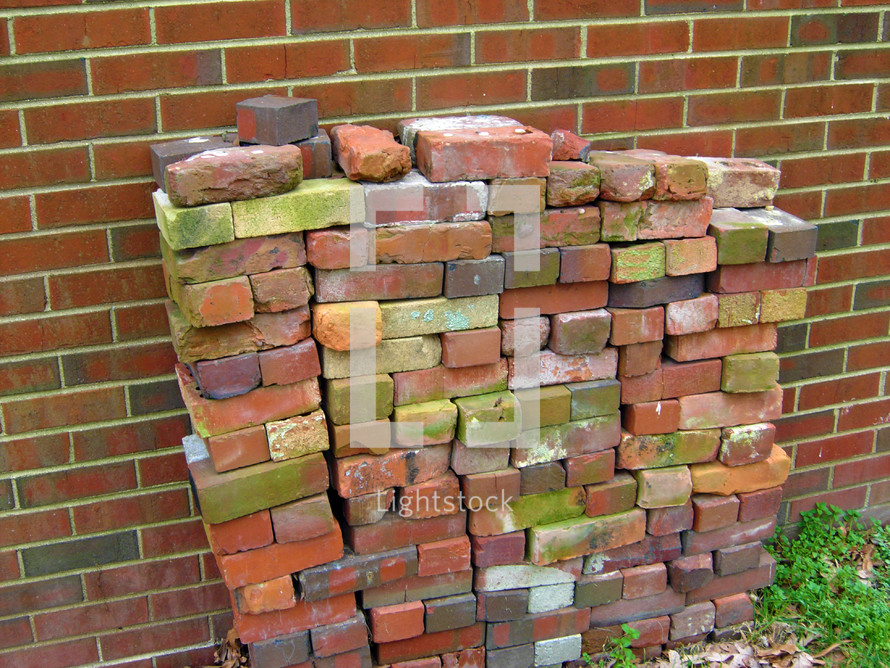 A pile of red stacked loose bricks stacked against a red brick wall outdoors waiting  to be used in construction of a new sidewalk or addition.  