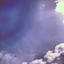 Plane flying across blue sky with cloud formations and sun beams.