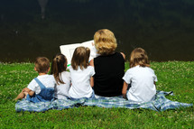Woman reading a book to children, sitting on the grass on a blanket