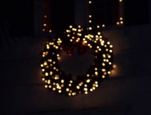 Christmas lights on a wreath - A Christmas Wreath decorated in Christmas white lights and red ornaments lights up the night in the background giving the night light to spread the joy of Christmas.
