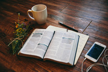 open Bible, journal, coffee mug, iPhone, earbuds, and flowers on a table 