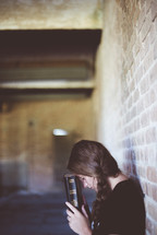 a woman leaning against a brick wall reading a Bible and praying 