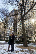 Embraced couple standing in the snow by a tree in front of buildings.