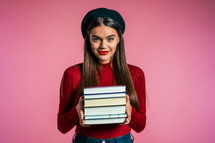 Pretty student on pink background in studio holds stack of university books from library. Girl in hat smiles, she is happy to graduate