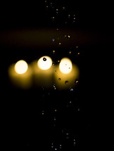 Three lit white candles behind pane of glass with rain drops.