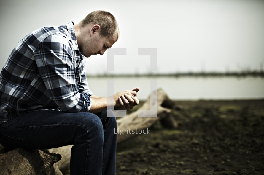A young man prays on a log next to a lake bed.