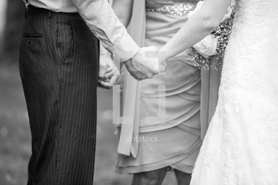 bride, groom, and bridesmaid holding hands in prayer