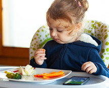Toddler eats in the high chair while watching movies on the mobile phone. 