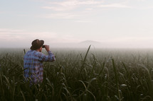 a man with binoculars standing in a field of tall green grass at dawn 