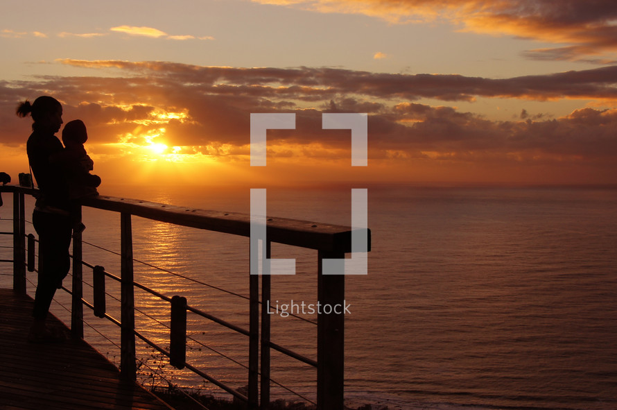 woman holding baby standing on pier with sunset over the water