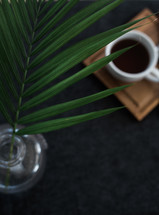 A sprig of green leaves in a vase next to a cup of coffee on a wooden board.