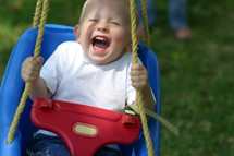 Boy toddler expressing pure chldlike joy as he swings from a tree in his yard with a parent in the far background watching. 