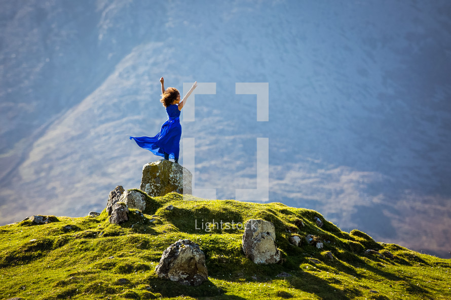 a girl in a blue dress standing on a rock on a green mountain with arms raised