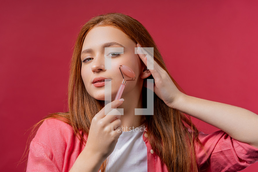 Young woman with rose quartz stone roller on pink background. Facial self care, beauty rituals, cosmetology, anti aging and anti-wrinkle treatment. High quality photo