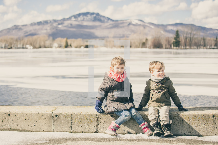 a boy and girl sitting on a curb in front of a frozen lake 