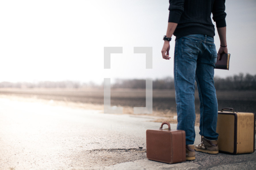 man standing next to a suitcase holding a Bible looking down a road wondering where to go from here