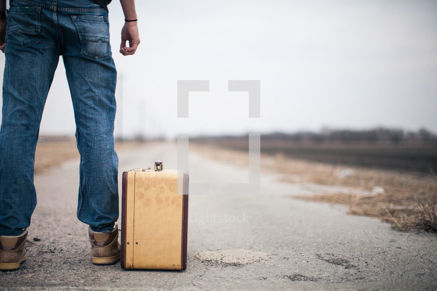 man looking down a road standing next to a suitcase