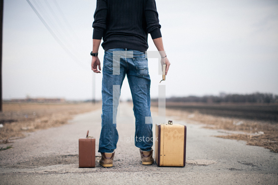 man standing next to suitcases holding a Bible looking down a long road and wondering where do I go from here