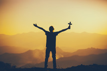 silhouette of a person with outstretched arms holding a cross standing on a mountaintop 