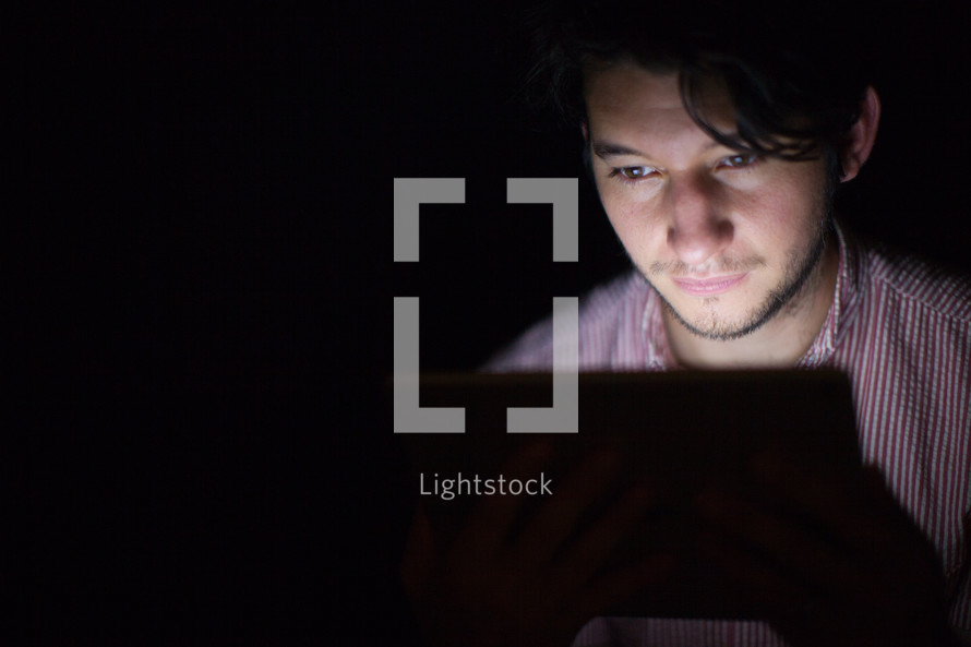 A man's face illuminated by the light from an electronic tablet.       
