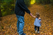 a father and toddler daughter walking holding hands through a fall forest 