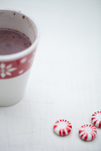 A cup of hot chocolate in a Christmas cup and peppermint candies on a white table.