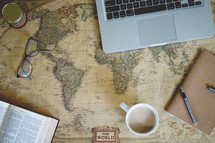 open Bible, mug, reading glasses, laptop,  pen, journal, and ink on a world map 