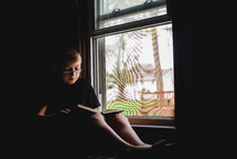 a boy reading a book in front of a window 