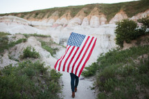 a woman walking on a trail carrying an American flag on her back 