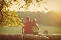 Couple sitting on hay bales in a pasture at sunset.
