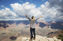woman standing at the edge of a canyon cliff with raised hands 