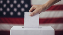 Close-up of a hand placing a ballot into a voting box with the American flag in the background.