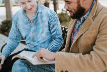 two men sitting on a bench reading a Bible and discussing scripture 