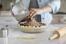 Hands pouring a topping onto a cherry pie.