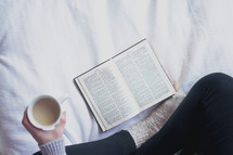 reading a Bible on a bed holding a mug 