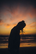 silhouette of a woman looking down standing on a beach at sunset 