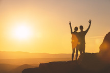 a couple with raised hands standing at the edge of a mountain at sunset 