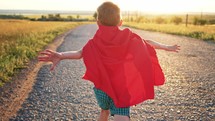 Boy Plays Superhero. Happy little boy are playing superhero. Kid run across road in red cloak at sunset time. Pretty child superhero hero in red cloak in nature. Freedom power concept.