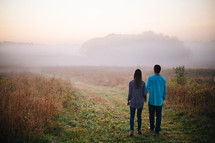 a young couple holding hands walking on a path through a field  