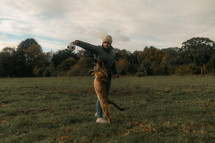 Dog jumping playing with owner in a field, young German Shepherd chasing dog toy, woman wearing tweed jacket and hat, winter, autumn